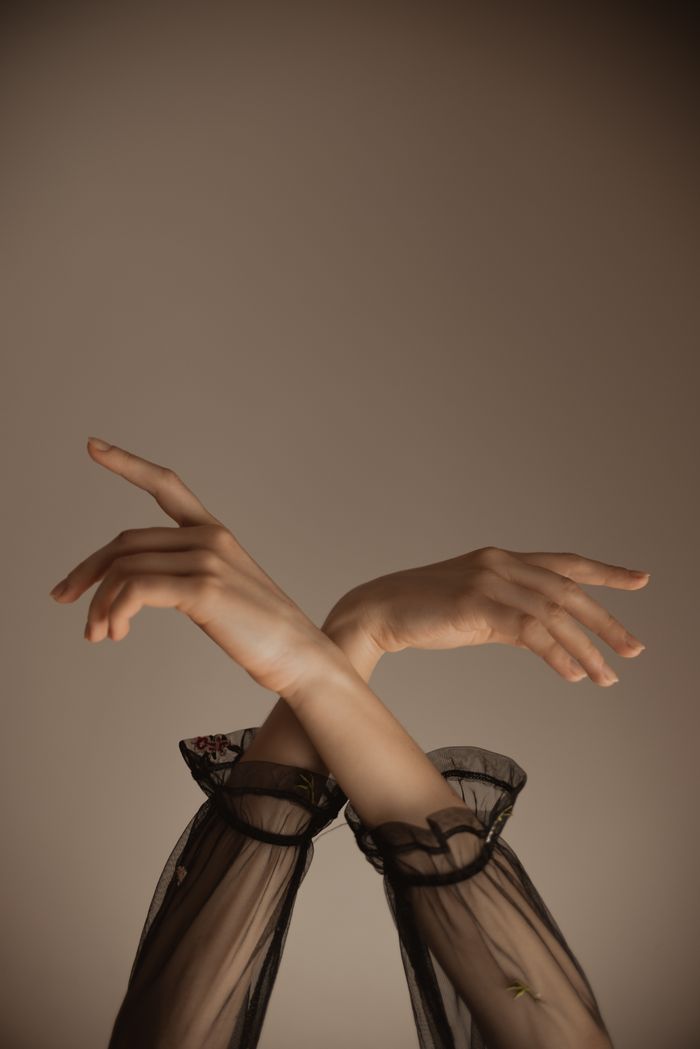 our-story Crop anonymous female dancer in transparent black dress keeping hands up while performing sensual dance movement against gray background