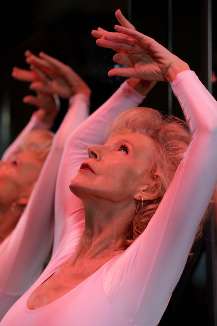 services-02 Senior woman in white leotard dancing elegantly with hands raised, reflected in a mirror.
